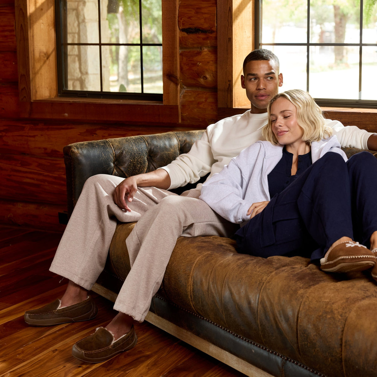 A man and a woman relax in a brown leather tufted sofa inside a rustic log cabin with wood floors and windows. The man wears a cream shirt and beige pants, and the woman wears a light blue cardigan, navy pants, and brown slip-on shoes.
#color_brown