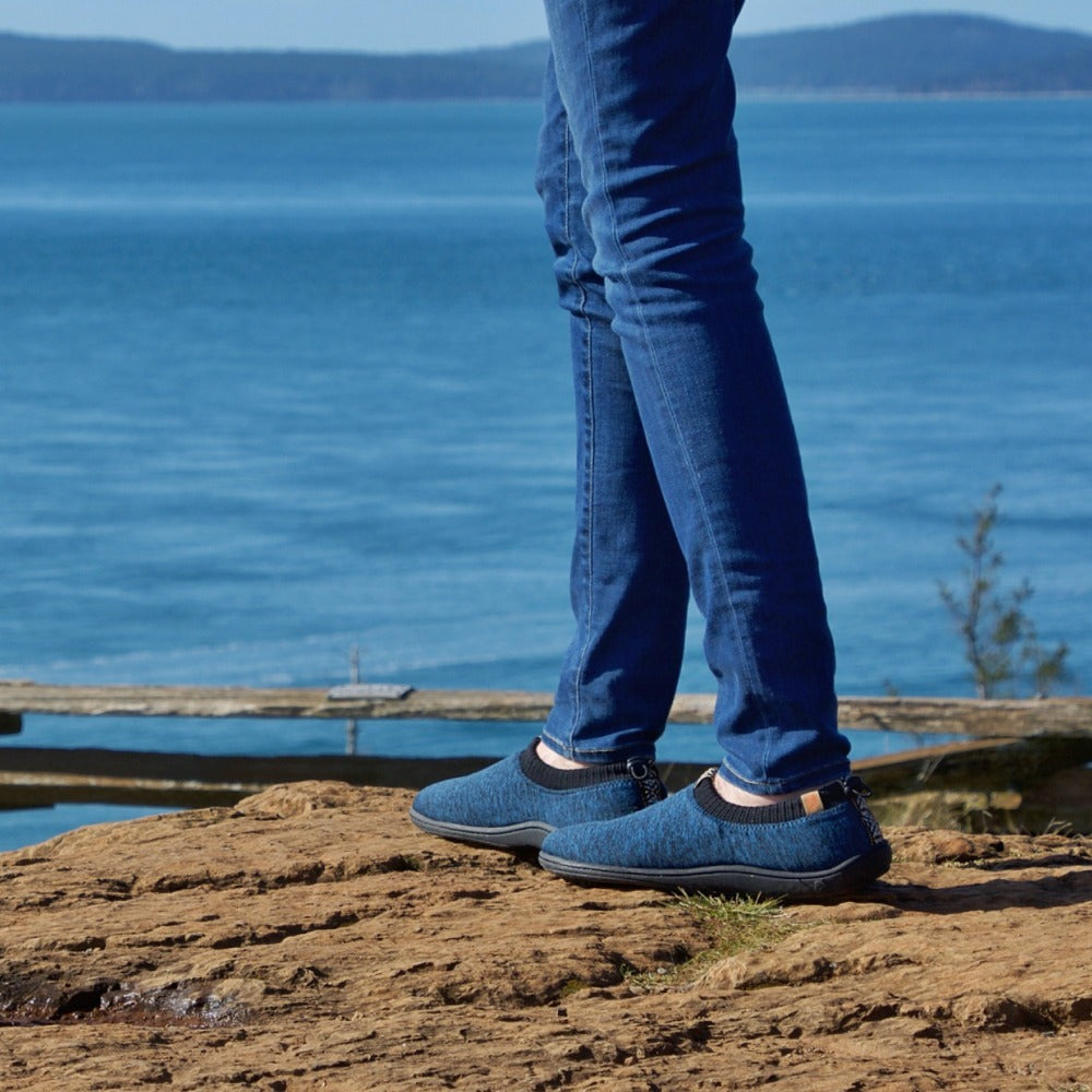 Men's Acorn Explorer Adventure Moc Male Model overlooking serene lake with mountains in the back on rocky terrain#color_navy-heather