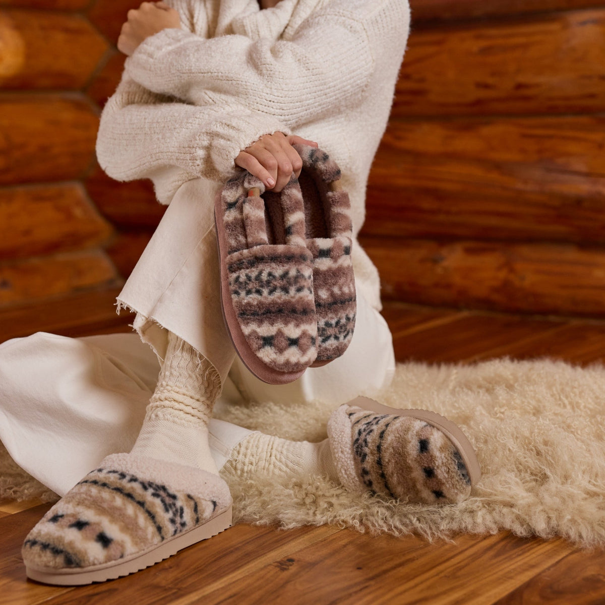 A person wrapped in a cream knit sweater sits on a fluffy cream rug in front of a rustic wood log wall, wearing cozy knit socks and slippers with a brown, beige, and gray geometric pattern. They hold one matching slipper in their hand.

#color_ivory