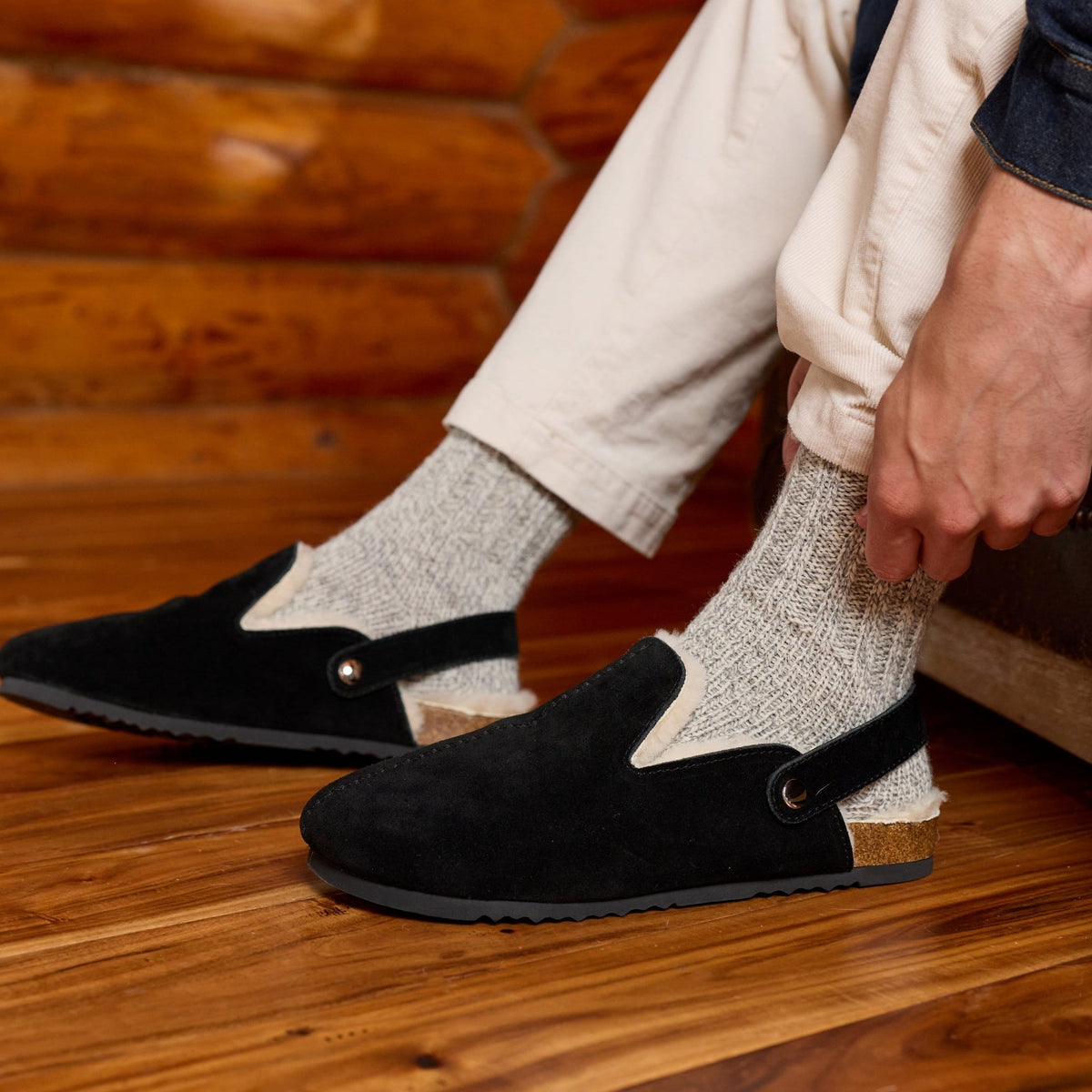 A person sitting in a rustic room wearing black slip-on slippers with elastic ankle straps, light gray knit socks, and cream pants.

#color_tan