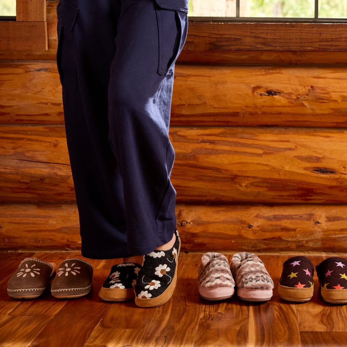 A person wearing dark navy pants stands on a wooden floor in front of a log cabin wall, with six pairs of slip-on mule slippers lined up below. The slippers are in various colors and patterns, including floral, knit, and star motifs.

#color_black-daisy